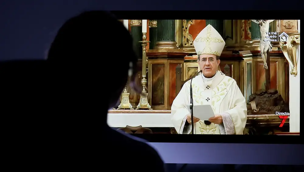 Una mujer ante una televisión con la imagen del arzobispo de Sevilla, Juan José Asenjo, durante la misa retransmitida desde la Catedral de la capital andaluza