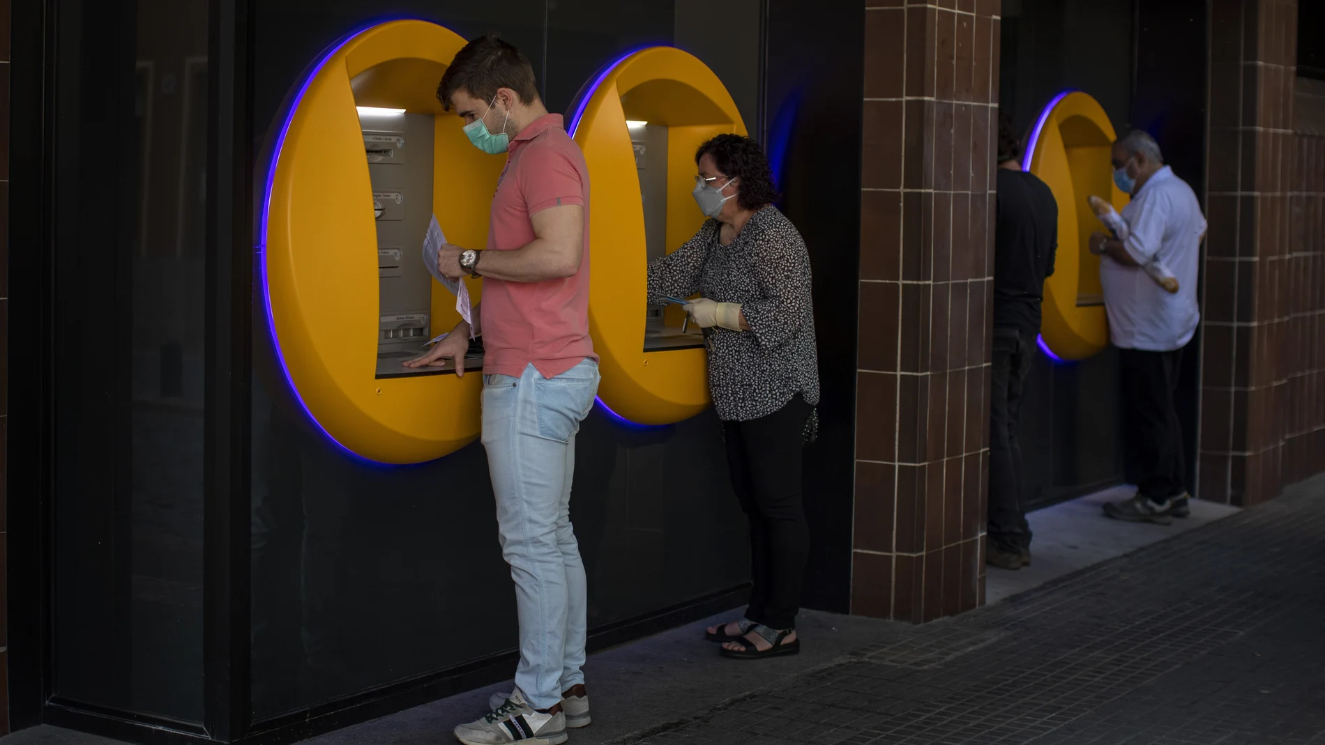 FILE - In this May 5, 2020, file photo, people wearing face masks use cash machines in Caldas de Montbui, near Barcelona, Spain. The coronavirus pandemic has reawakened debate about the continued viability of what has been the physical lifeblood of global economies: paper money and coins. (AP Photo/Emilio Morenatti)