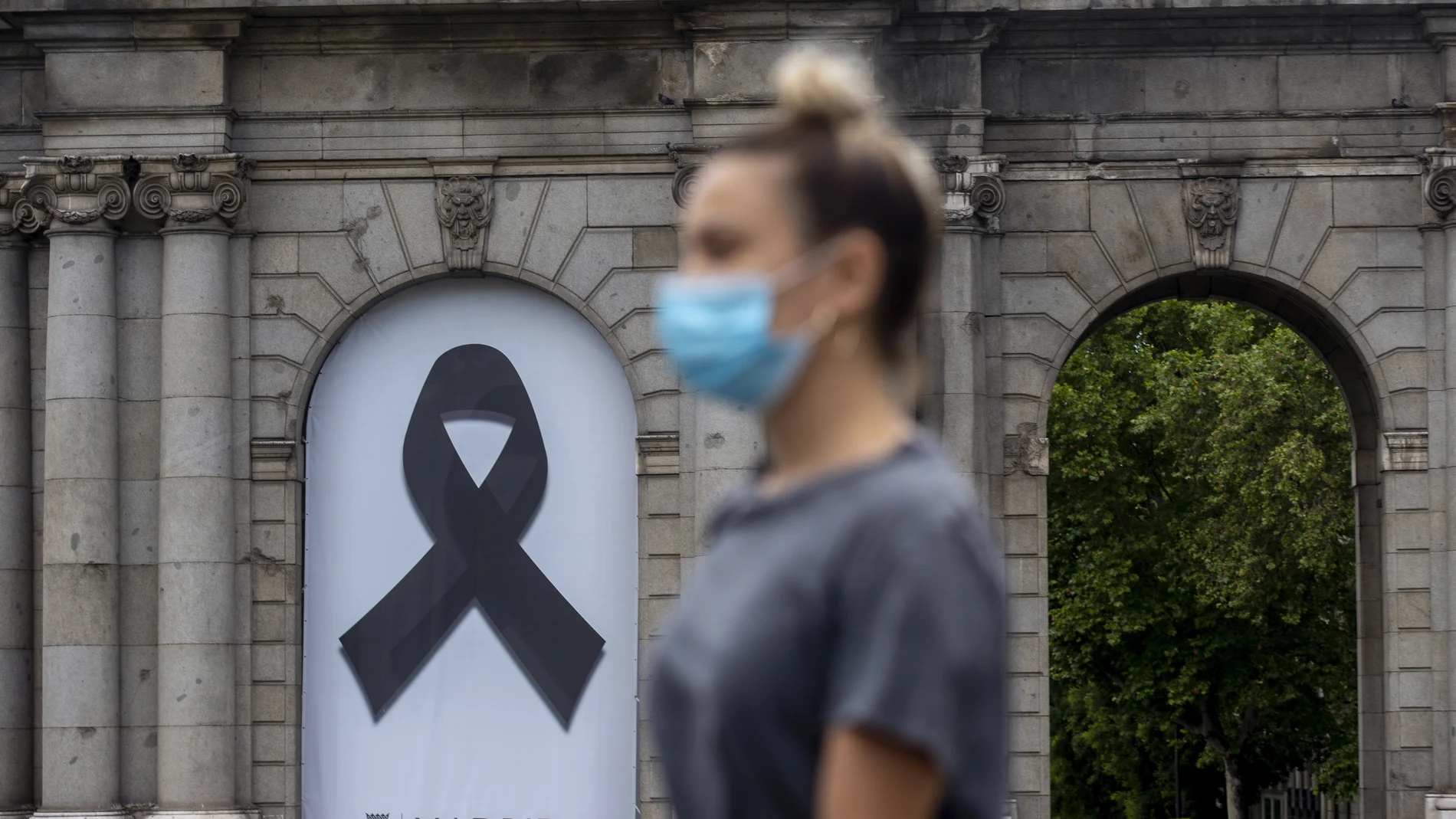 Una mujer con mascarilla frente a la Puerta de Alcalá donde un crespón negro recuerda a los fallecidos por coronavirus