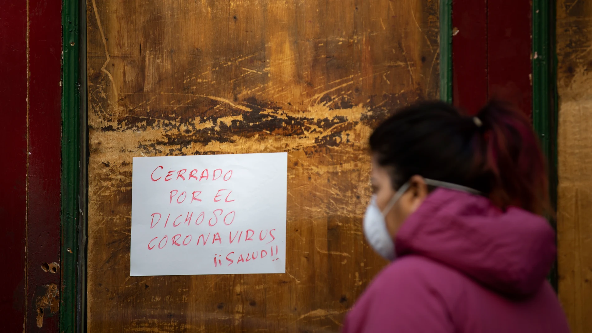 Imágen de una mujer pasando por delante de un cartel en un comercio que reza: "Cerrado por el dichoso coronavirus".