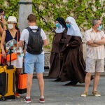 Unos turistas esperan el autobús mientras una pareja de monjas de la Orden de Santa Ángela de la Cruz pasan por detrás con mascarillas