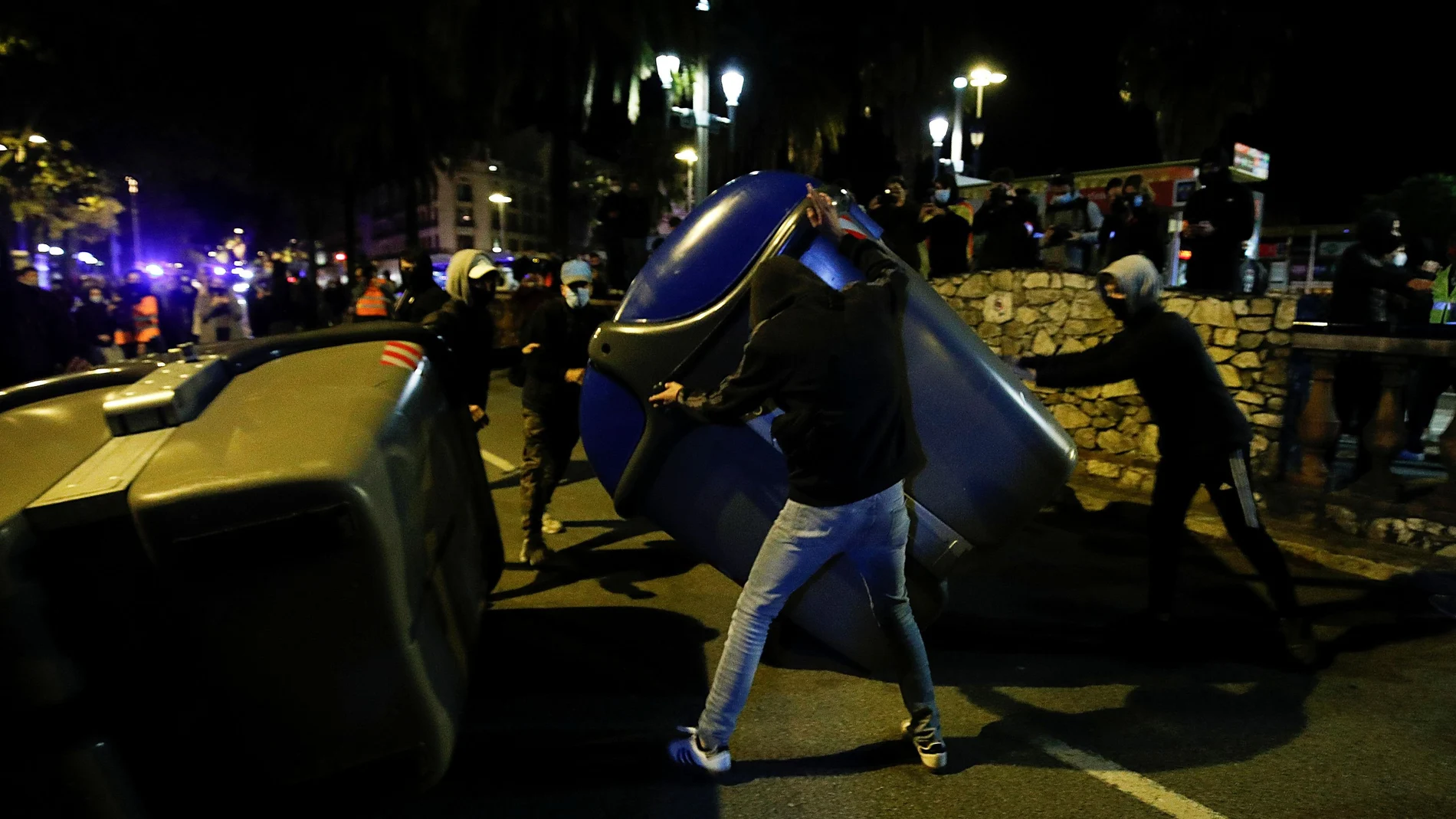 Varios manifestantes, de perfil muy joven, tumbando contenedores en el paseo de Colón de Barcelona