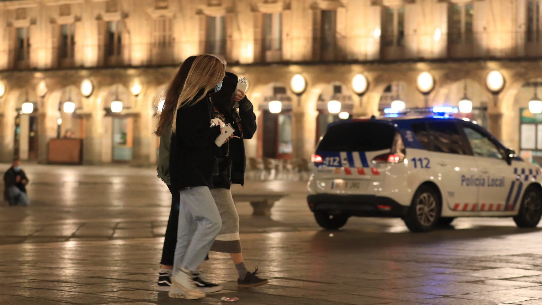 Unas jóvenes pasean por la Plaza Mayor de Salamanca junto a una coche de la Policía Nacional