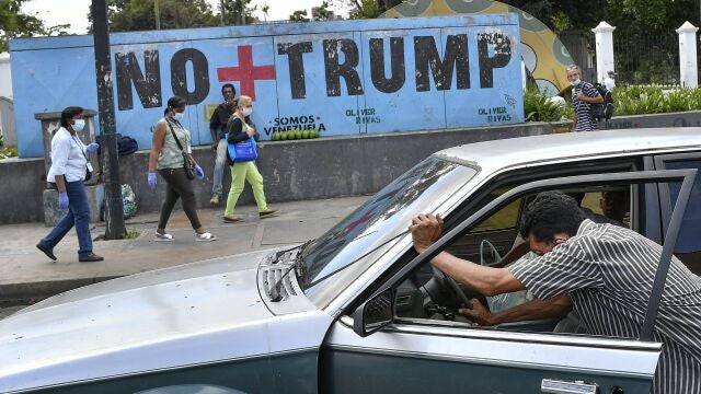 A man pushes his car that ran out of gas as people wearing protective face masks as a precaution against the spread of the new coronavirus, walk past a mural with a message that reads in Spanish: " No more Trump" in Caracas, Venezuela, Sunday, Nov. 8, 2020. Venezuelan President Nicolas Maduro has sent a congratulatory message to U.S. President-elect Joe Biden who won the U.S. presidential election, defeating incumbent Donald Trump. (AP Photo/Matias Delacroix)