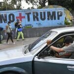 A man pushes his car that ran out of gas as people wearing protective face masks as a precaution against the spread of the new coronavirus, walk past a mural with a message that reads in Spanish: " No more Trump" in Caracas, Venezuela, Sunday, Nov. 8, 2020. Venezuelan President Nicolas Maduro has sent a congratulatory message to U.S. President-elect Joe Biden who won the U.S. presidential election, defeating incumbent Donald Trump. (AP Photo/Matias Delacroix)