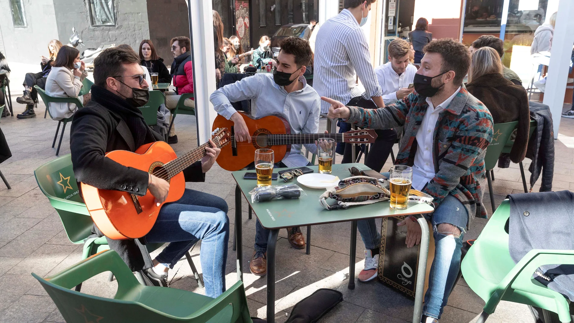 Tres jóvenes tocan la guitarra en la terraza de un bar de la Plaza de la Universidad de Murcia mientras toman el aperitivo