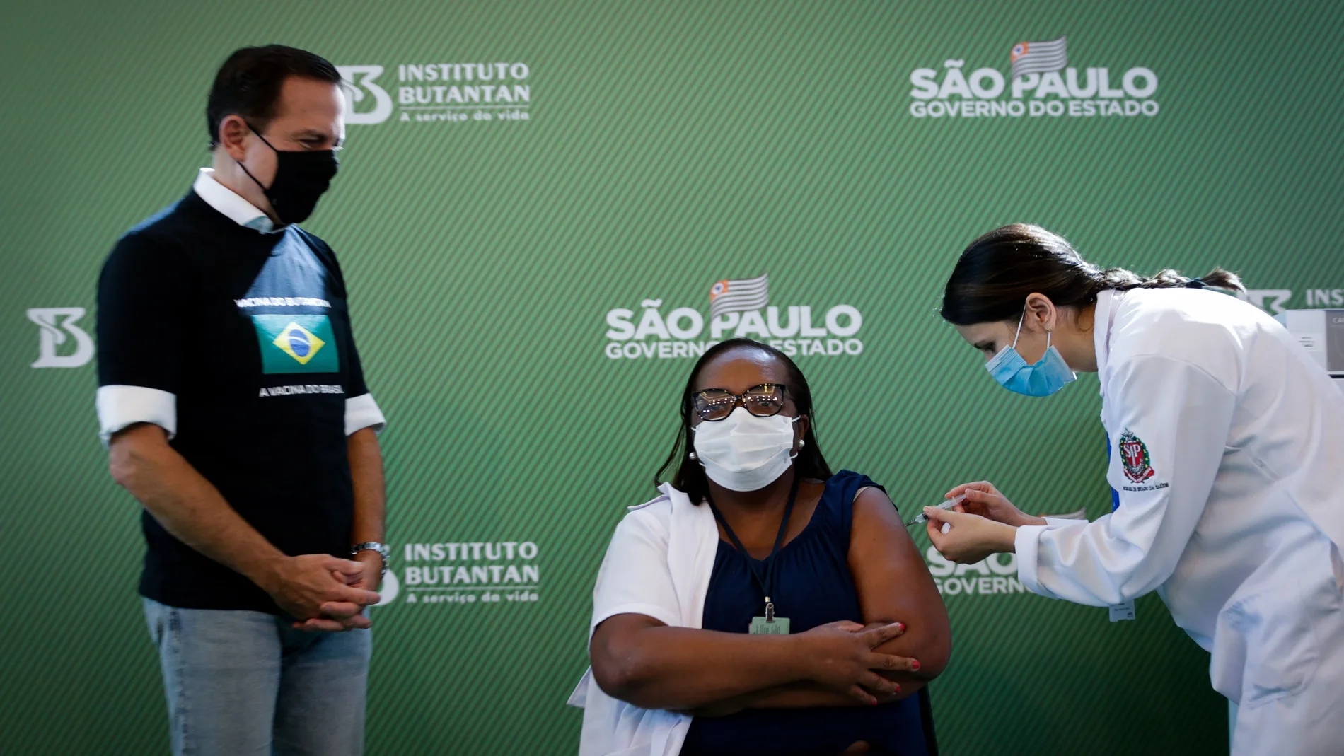 AME9923. SAO PAULO (BRASIL), 17/01/2021.- Mônica Calazans (c), enfermera del Hospital las Clínicas de la capital paulista recibe la vacuna contra la covid-19 en presencia del gobernador de Sao Paulo, Joao Doria (i), hoy, en Sao Paulo (Brasil). Brasil, uno de los países del mundo más afectados por la pandemia del coronavirus, aplicó este domingo en Sao Paulo la primera dosis de la vacuna contra la covid-19 a la enfermera de 54 años, en momentos en los que el gigante latinoamericano enfrenta una segunda ola de la enfermedad. La primera dosis de la vacuna desarrollada por el laboratorio chino Sinovac y el Instituto brasileño Butantan fue aplicada tan solo minutos después de la aprobación de su uso de emergencia por parte de la Agencia Nacional de Vigilancia Sanitaria (Anvisa). EFE/ Fernando Bizerra Jr