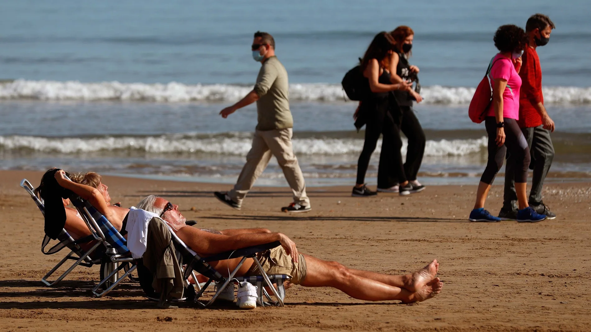 Dos personas toman el sol en la Playa de la Malvarrosa (Comunidad Valenciana) mientras otras pasean por la orilla