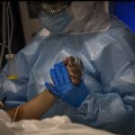 Un sanitario de la UCI ataviado con un EPI, mascarilla y guantes de látex toma la mano de un paciente de coronavirus
