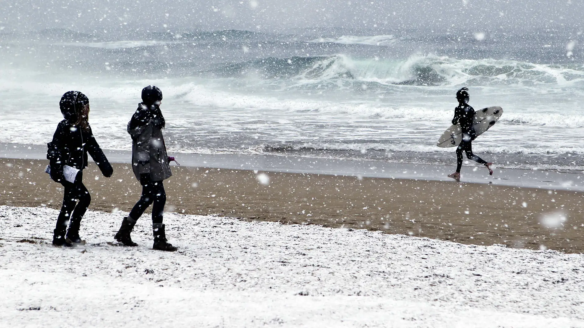 Dos chicas observan a un surfista momentos antes de meterse al agua, hoy en la playa de La Zurriola de San Sebastián, bajo una intensa nevada. EFE/Javier Etxezarreta