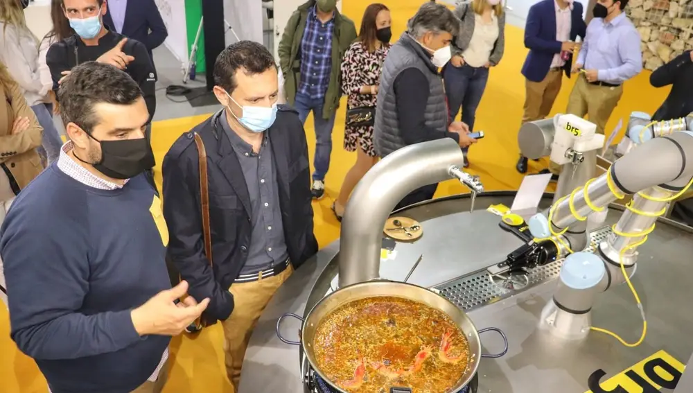 GRAF3761. MÁLAGA, 28/04/2021.- La primera paella creada por un robot, capaz de ser preparada sin necesidad de supervisión de un cocinero, llega para revolucionar este plato típico español y el propio sector de la restauración, ante el que se ha presentado en el Salón de Innovación en Hostelería H&T, en Málaga. En la imagen, varias personas junto al director general de br5, Enrique Lillo (izq), observan cómo actúa el brazo robotizado en la paella. EFE/ Daniel Luque