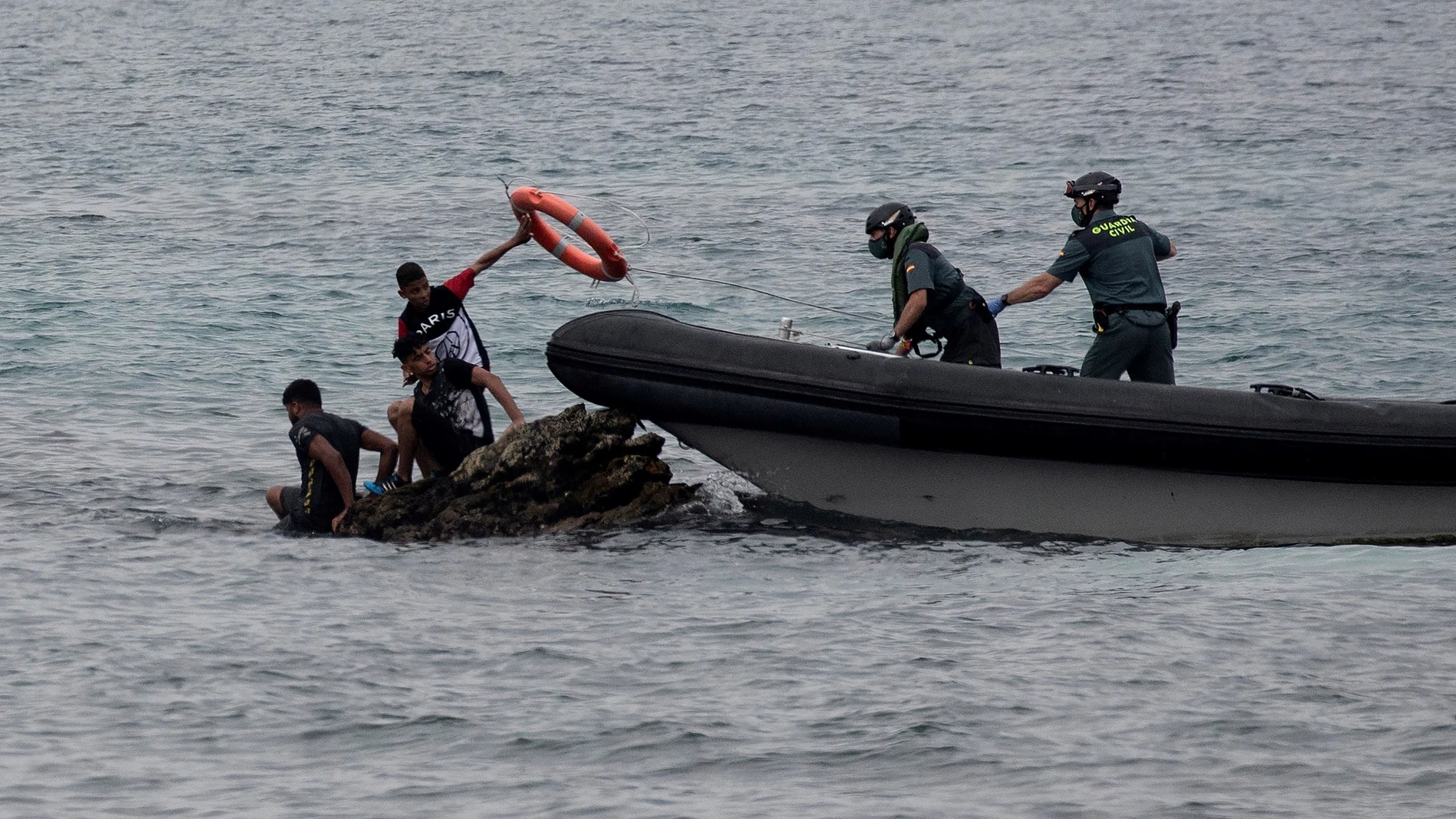 Miembros de la Guardia Civil lanzan un flotador a varios menores que esperan encima de unas rocas en la playa ceutí de El Tarajal
