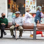 Imagen de dos señores mayores en un banco sentados en el que se puede ver a uno de ellos con mascarilla y el otro sin ella con un paraguas y un bastón en la mano. Imagen de tercera edad y jubilados.