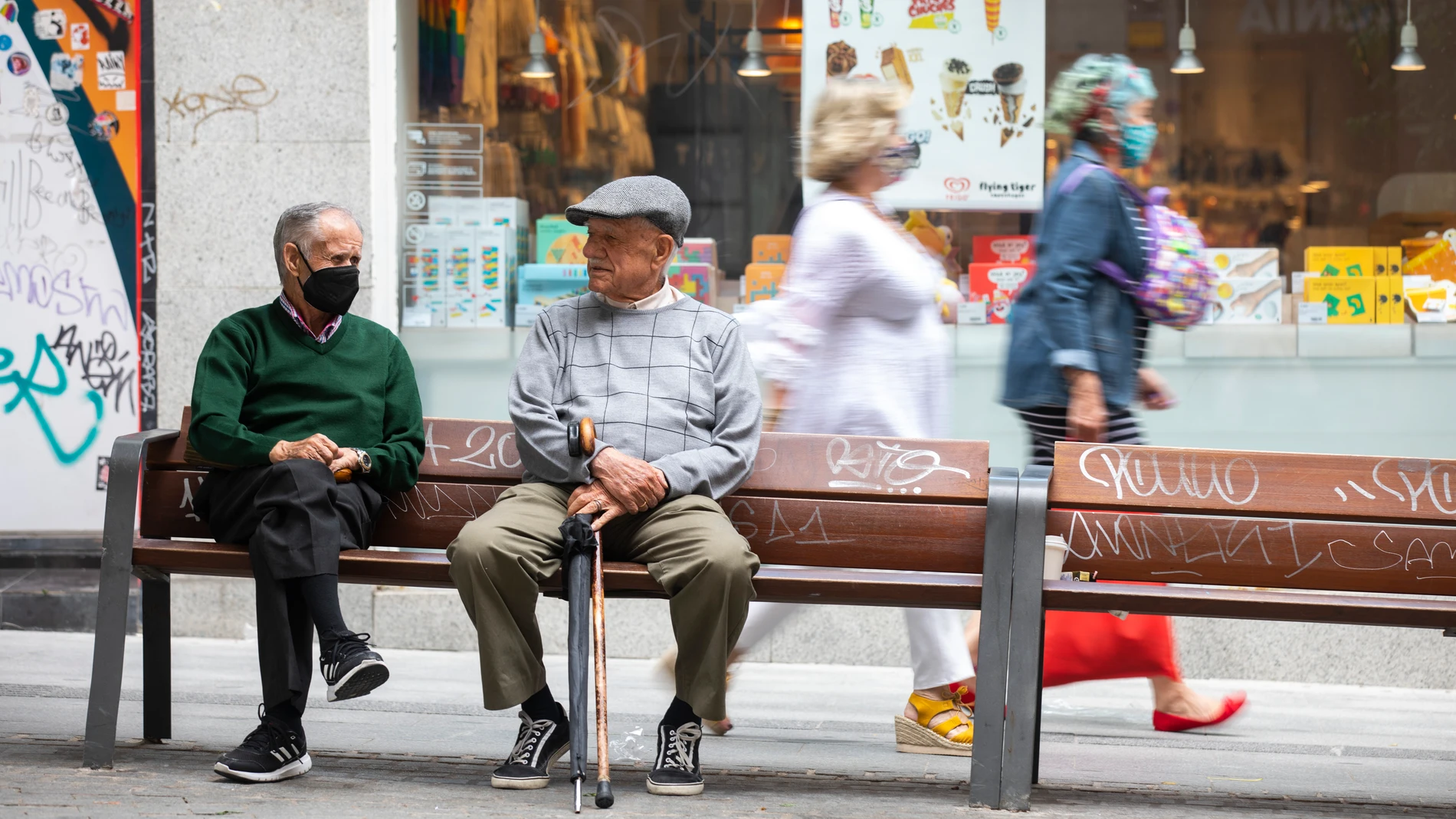 Imagen de dos señores mayores en un banco sentados en el que se puede ver a uno de ellos con mascarilla y el otro sin ella con un paraguas y un bastón en la mano. Imagen de tercera edad y jubilados.