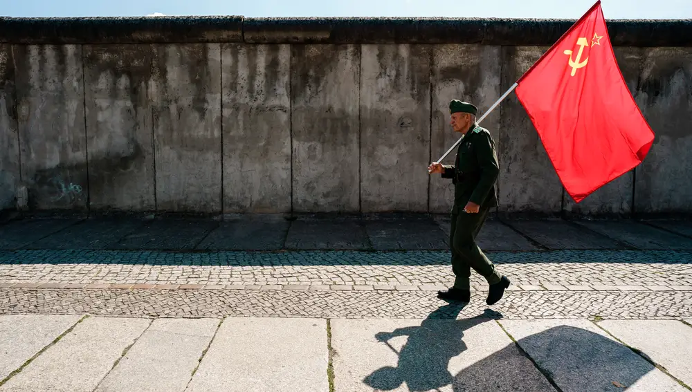 Un hombre con uniforme del ejército soviético lleva una bandera soviética junto a los restos conservados del Muro de Berlín tras la ceremonia de conmemoración del 60 aniversario de la construcción del Muro de Berlín en Berlín, Alemania, 13 de agosto de 2021. El 13 de agosto de 1961 comenzó la construcción del Muro de Berlín. (Alemania) EFE/EPA/CLEMENS BILAN