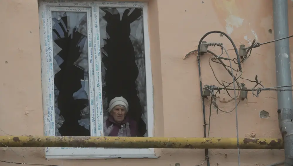 Una mujer mayor a través de una ventana rota de un edificio residencial, dañado como resultado de un bombardeo, en Donetsk, República Popular de Donetsk | Fuente: Ilya Pitalev / Sputnik