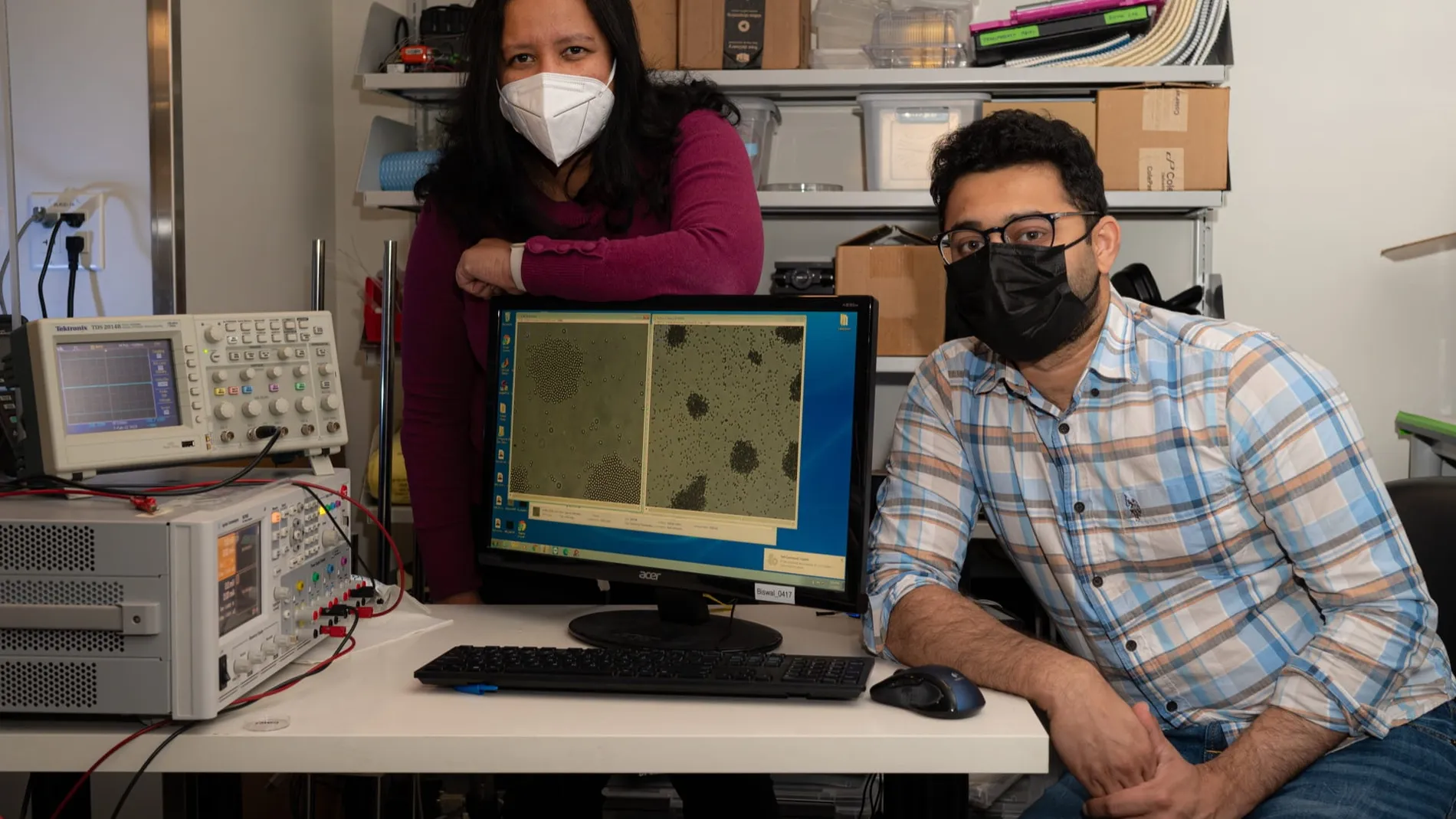 Sibani Lisa Biwal y Kedar Joshi en su laboratorio con una imagen de las partículas magnéticas mostrada en el ordenador. A la izquierda hay aparatos de laboratorio.
