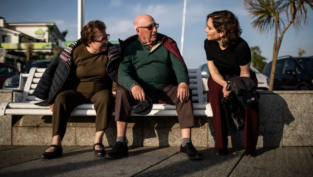 1. Ayuso conversa con un matrimonio en un banco del Paseo del Muro de San Lorenzo de Gijón