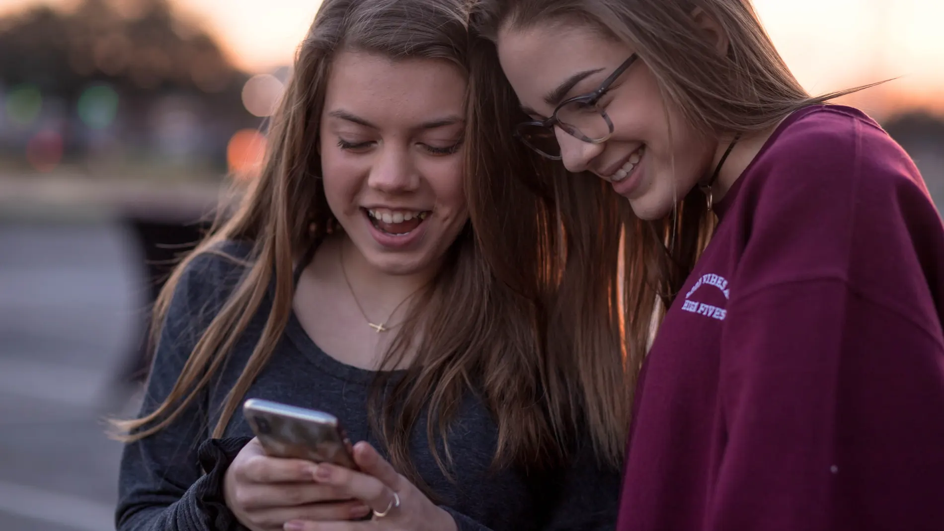 Dos chicas jóvenes, de pie con las cabezas juntas mirando el móvil de una de ellas y riéndose