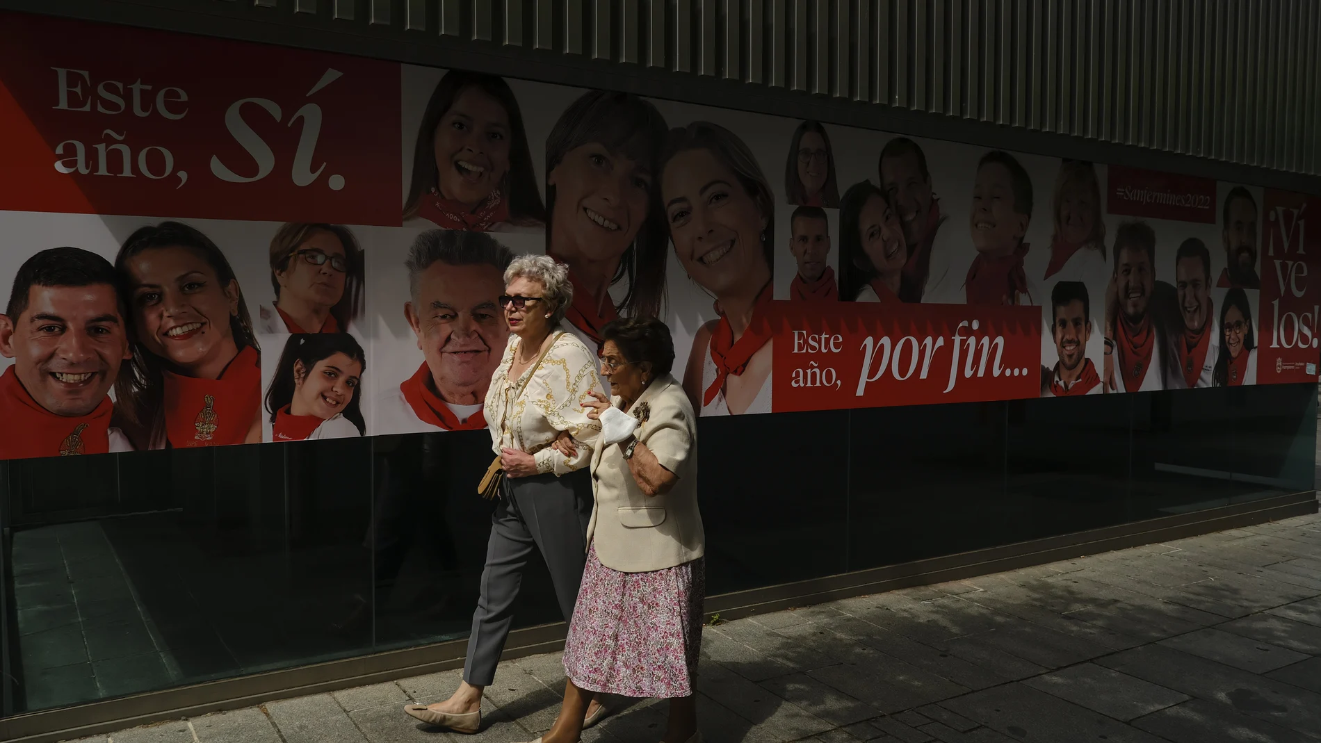 Personas paseando por Pamplona ante un cartel que anuncia los San Fermines, que comienzan el jueves, después de dos años sin celebrarse por la pandemia de covid