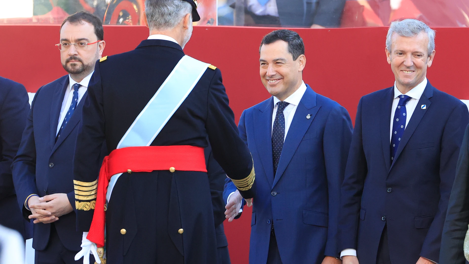 El rey saluda al presidente de Andalucía, Juanma Moreno (2d), en presencia del presidente de Asturias, Adrián Barbón (i), y el presidente de Galicia, Alfonso Rueda (d), al inicio del desfile del Día de la Fiesta Nacional, este miércoles, en Madrid. EFE/Zipi Aragón