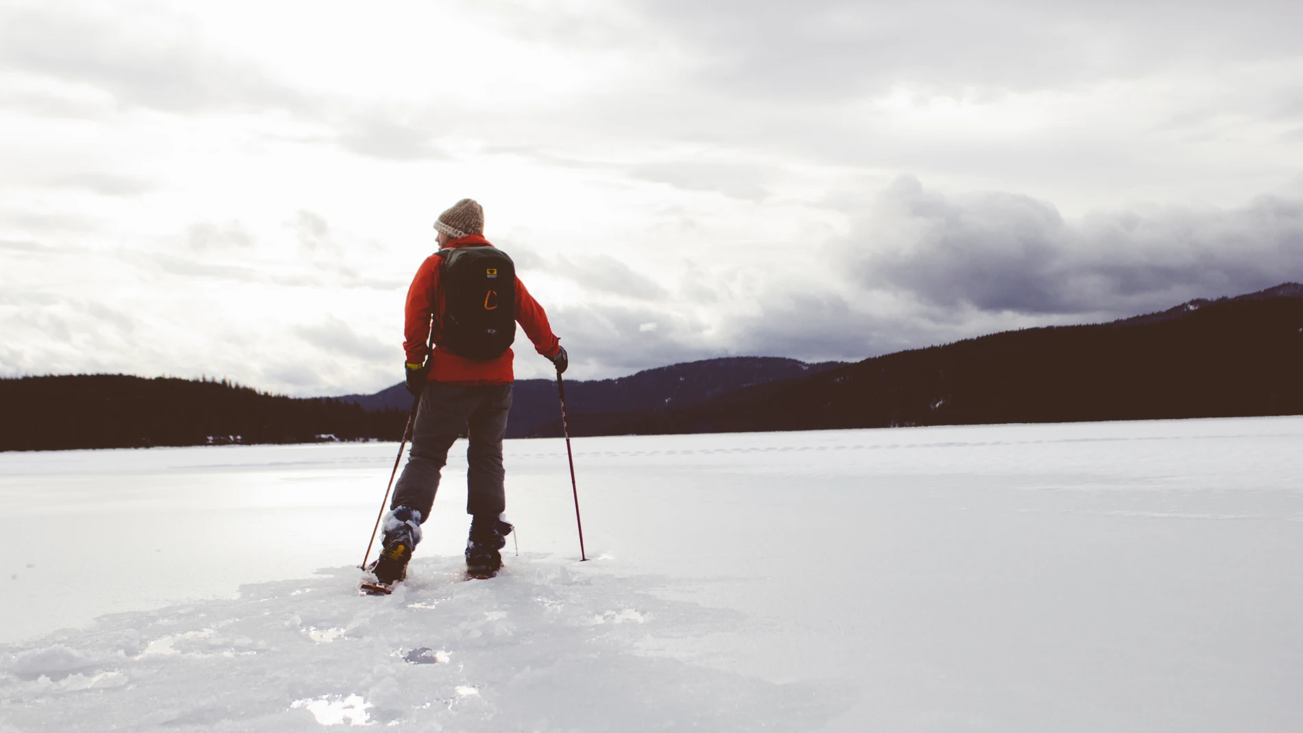 Un hombre con abrigo, botas, mochila y bastones, de espaldas a la cámara sobre una gran masa de hielo con montañas al fondo