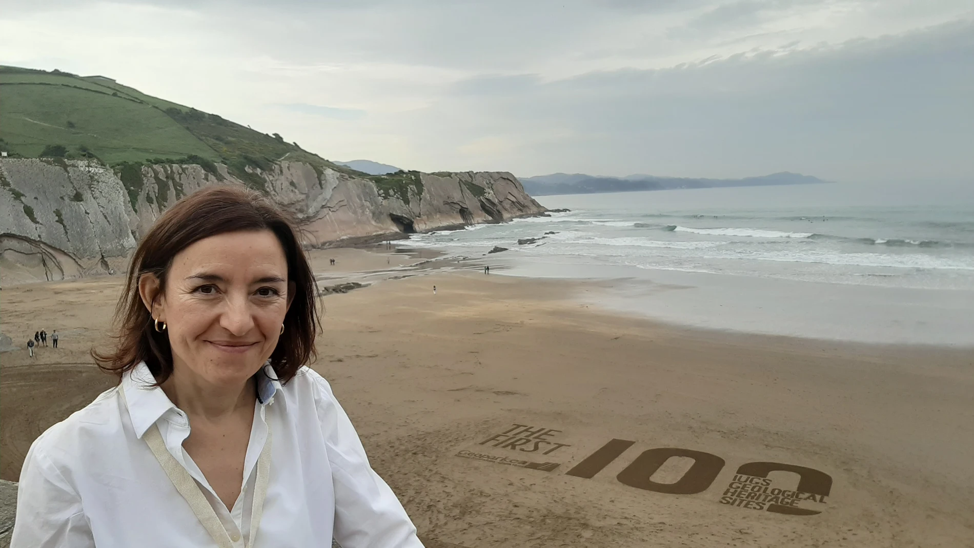 Una mujer con camisa blanca sonriendo delante de una playa donde se ve el número 100 pintado en la arena