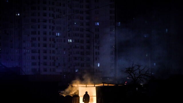 KUn hombre cerca de basura ardiendo frente a un edificio de apartamentos durante un corte de electricidad programado en Kyiv. Detrás, un edificio de apartamentos sin luz