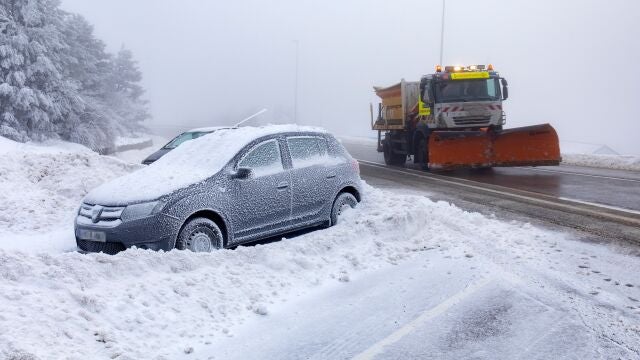 Un coche cubierto de nieve y una máquina quitanieves en la carretera de acceso al Puerto de Navacerrada