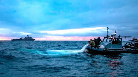 El buque de guerra anfibia de la clase Harpers Ferry USS Carter Hall navega en el fondo mientras los marineros de la Armada recuperan una parte del sobre del globo espía chino.