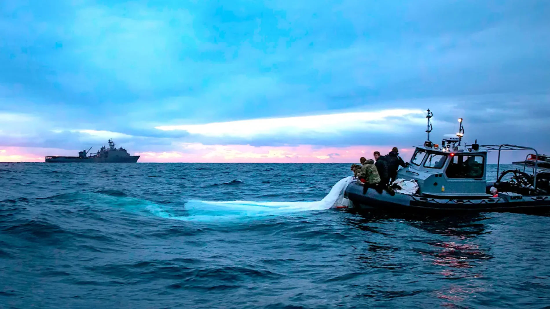 El buque de guerra anfibia de la clase Harpers Ferry USS Carter Hall navega en el fondo mientras los marineros de la Armada recuperan una parte del sobre del globo espía chino.