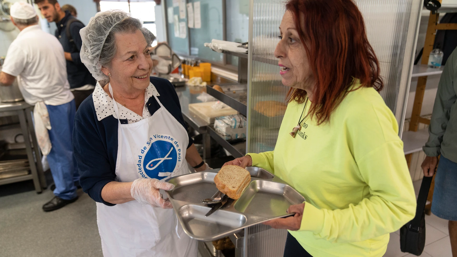 María Paz, voluntaria de 79 años, da la bandeja para la comida a Silvia, canaria de 59 años, en el comedor social Santiago Masarnau que cumple 25 años