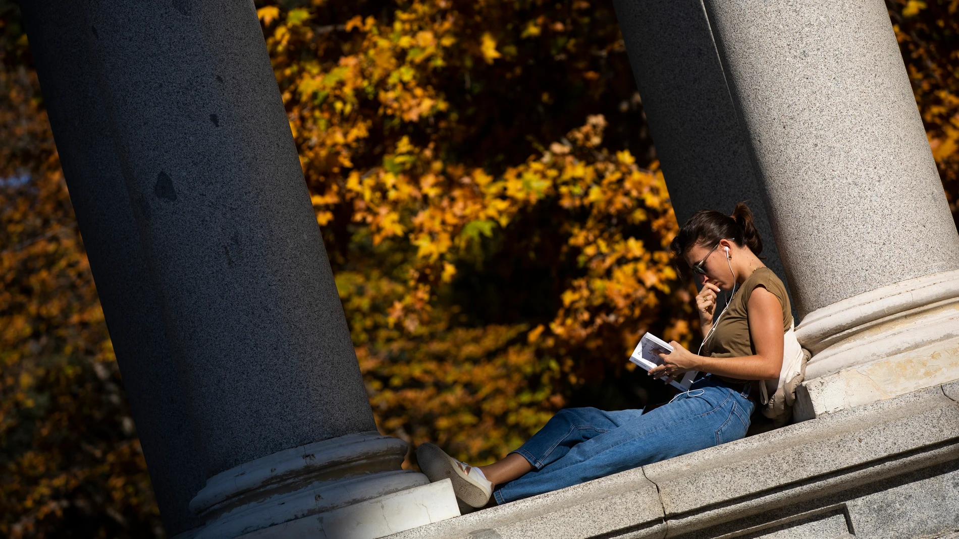 Otoño en el Parque del Retiro. Día de altas temperaturas y buen tiempo a pesar de las fechas. Una joven lee un libro y escucha música al sol. © Jesús G. Feria.