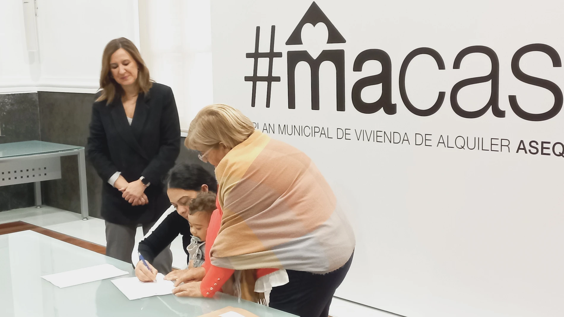 Una madre con su bebé en brazos firma el contrato de alquiler en presencia de la alcaldesa