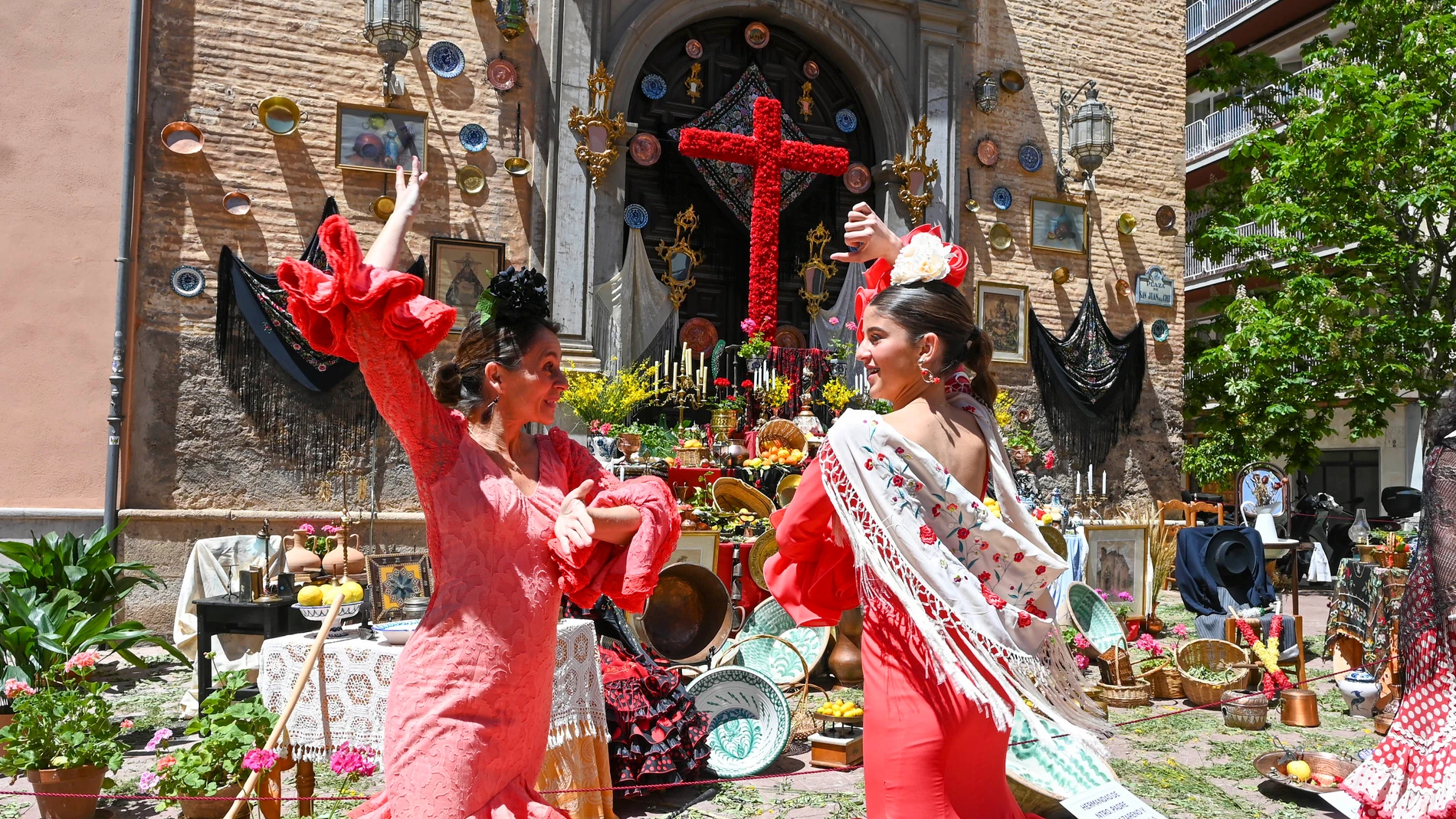 GRANADA, 03/05/2024.- Unas jóvenes bailan junto a una de las cruces que adornan las principales plazas y barrios de Granada que este viernes exhiben adornadas con flores, mantones y cerámicas con motivo del tradicional Día de la Cruz, con dos días más de celebración pero sin barras en las calles, solo en recintos cerrados. EFE/Miguel Ángel Molina