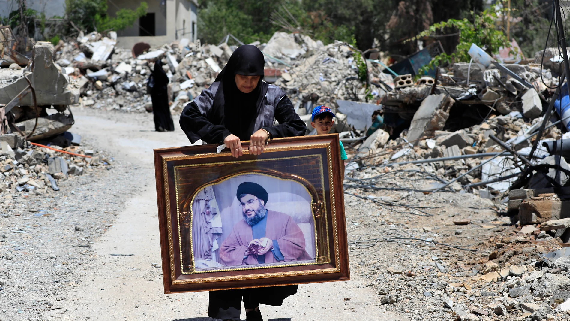 FILE - A woman carries a frame that shows a portrait for Hezbollah leader Sayyid Hassan Nasrallah, as she passes by destroyed houses that were hit by Israeli airstrikes, in Aita al-Shaab, a Lebanese border village with Israel, south Lebanon, June 29, 2024. (AP Photo/Mohammed Zaatari, File)