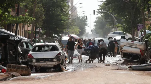 VALENCIA, 30/10/2024.- Unas personas caminan por una calle cubierta de lodo y llena de coches amontonados tras las intensas lluvias por la fuerte DANA hoy en Valencia