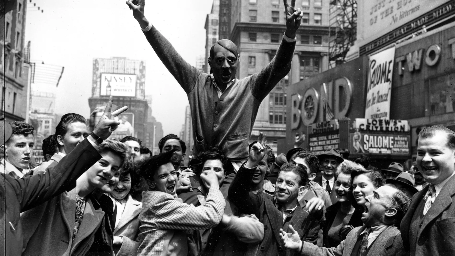 FILE - New Yorkers gather around a youth wearing a mask of Adolf Hitler to cheer the news of the surrender of Germany in New York's Times Square on May 7, 1945. (AP Photo/Harry Harris, File)