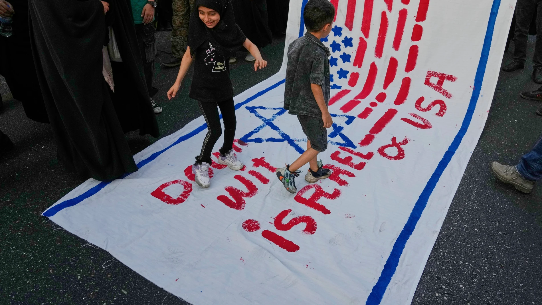 Two children walk on an anti-U.S. and anti-Israeli banner during a rally marking the Muslim Shiite holiday of Eid al-Ghadir, which commemorates the Prophet Muhammad naming his successor, Ali, who is revered as the first Shiite imam, in Tehran, Iran, Saturday, June 14, 2025.(AP Photo/Vahid Salemi)