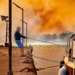 A man ties a boat at the port of Lithi in Chios, while a wildfire burns through forested land, Greece, 24 June 2025 (issued 25 June 2025). Greek authorities on 25 June announced the arrest of a 30-year-old Georgian woman suspected of tossing away a lit cigarette that helped start one of the wildfires that broke out between 22 and 23 June, while investigations are still underway to determine the cause of the other blazes on the island. 