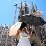 Una turista utiliza dos sombrillas para protegerse del sol frente a la Sagrada Familia en Barcelona