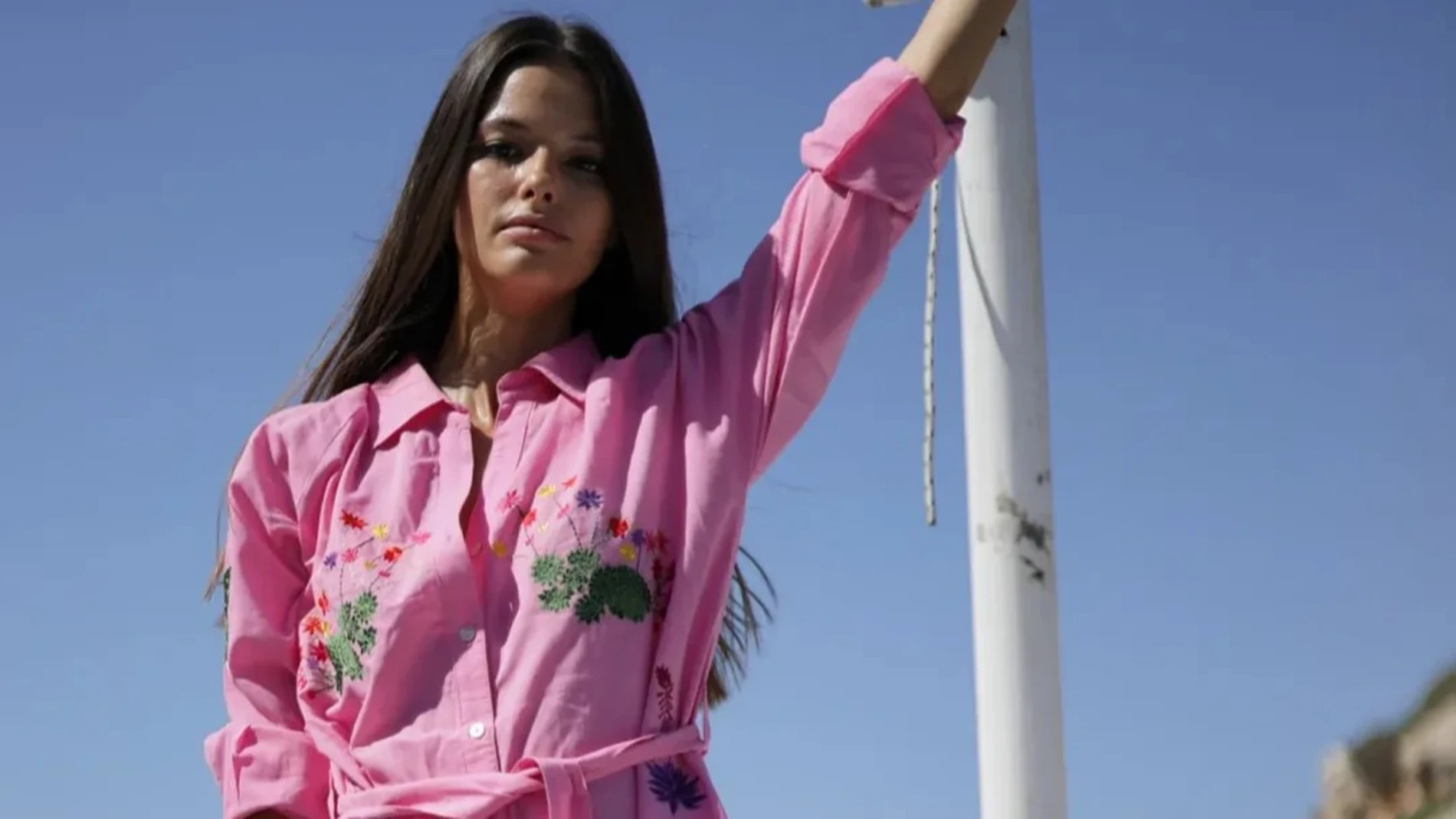 Mujer con vestido camisero rosa bordado y cinturón, de Luna Llena, bajo el sol en un entorno costero.