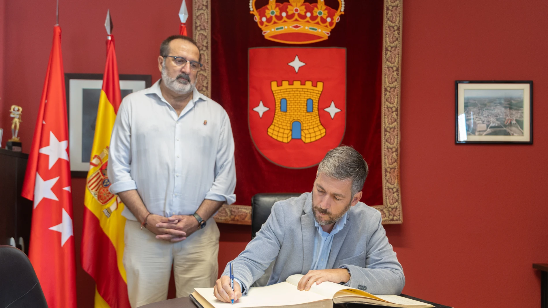 El consejero de Presidencia, Justicia y Administración Local de la Comunidad de Madrid, Miguel Ángel García Martín (d), firma el Libro de Honor del Ayuntamiento de Torrejón de Velasco, durante su visita a una de las cuatro oficinas móviles de Atención al Ciudadano, en la Plaza de España, a 8 de agosto de 2025, en Torrejón de Velasco, Madrid (España). García Martín ha visitado la oficina para hacer balance de este servicio público que el Gobierno regional pone a disposición de los vecinos de p...