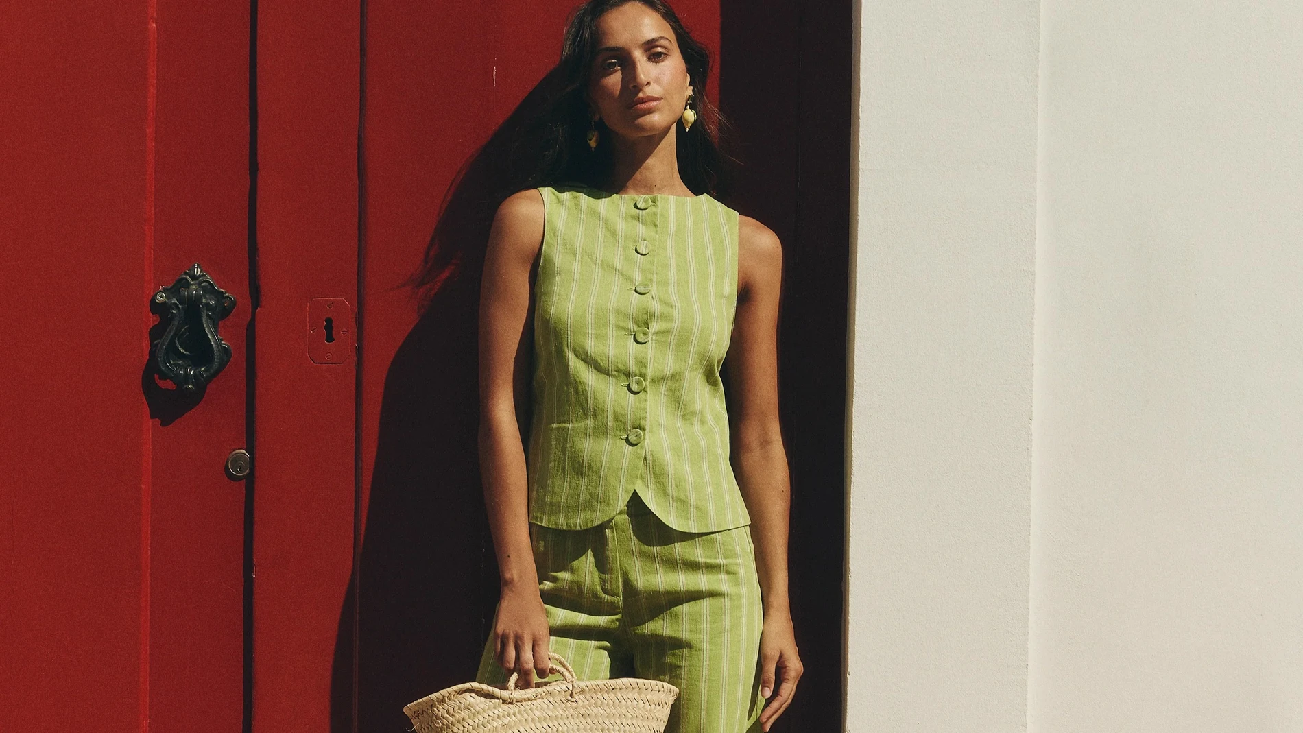 Conjunto de lino con rayas verdes y amarillas de Baymo, combinado con capazo de palma natural, en un look de verano fotografiado frente a una puerta roja.