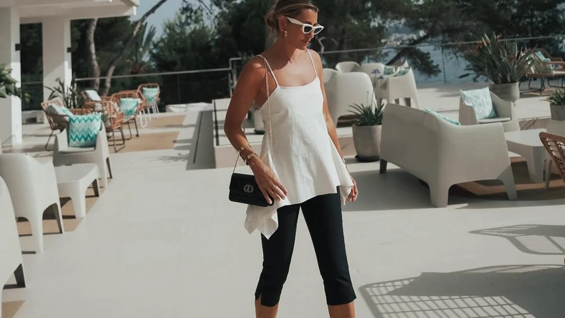 Influencer con pantalón capri negro, top fluido blanco de tirantes, bolso negro de cadena y flip flop de tacón bajo, caminando en una terraza con mobiliario de exterior.
